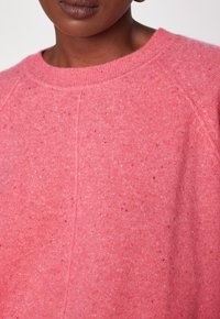Primo piano di una persona che indossa un maglione a maglia testurizzata rosa con scollo rotondo a coste e dettagli di cucitura visibili.