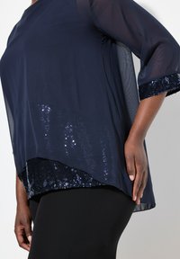 Navyblaues Oberteil mit durchsichtigen Ärmeln, verziert mit Pailletten am Saum und an den Bündchen. Darunter sind angepasste schwarze Hosen teilweise sichtbar.