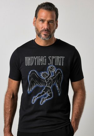 Mężczyzna z krótkimi włosami i brodą, ubrany w czarną koszulkę z grafiką skrzydlatego koszykarza i tekstem "UNDYING SPIRIT" na przodzie.