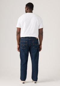 Mann, der mit dem Rücken zur Kamera steht, trägt ein weißes T-Shirt, dunkelblaue Jeans mit hinteren Taschen und weiße Sneakers vor einem einfarbigen Hintergrund.