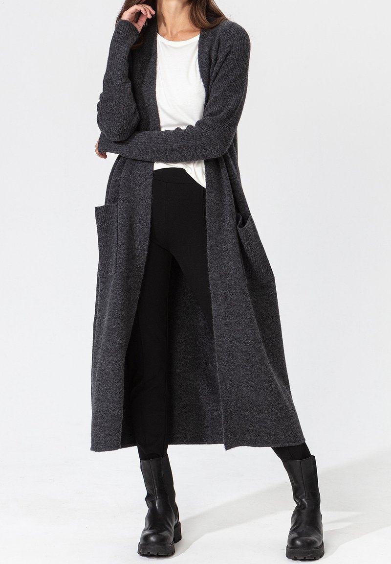 Long gilet gris en maille texturée avec devant ouvert, manches longues et poches latérales, porté sur une chemise blanche et un pantalon noir.