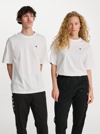 Due persone indossano t-shirt bianche di cotone coordinate con un piccolo logo nero. Le abbinano a pantaloni neri con accenti laterali a fantasia.