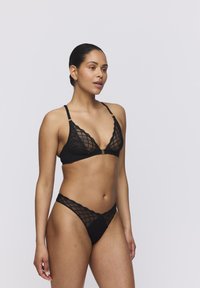 Conjunto de lencería de encaje negro que incluye un bralette triangular con un patrón entrecruzado y un tanga a juego con tirantes finos. Acentos en hardware dorado.