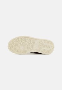 Suola di una scarpa realizzata in un materiale gommoso beige chiaro con un motivo di presa testurizzato e un accento circolare, contornata da un bordo scuro.