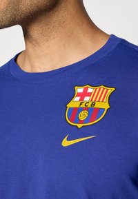 Kraljevsko plava kratkotrajna majica s kratkim rukavima sadrži žuti Nike logo i šareni grb FC Barcelone s crvenim i plavim prugama te zlatnim akcentima.
