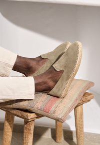 Hellolivgrüne Slipper aus Wildleder mit runder Zehenkappe, Jutensohle und sichtbaren Nähten, getragen beim Ausruhen auf einem gewebten Kissen.