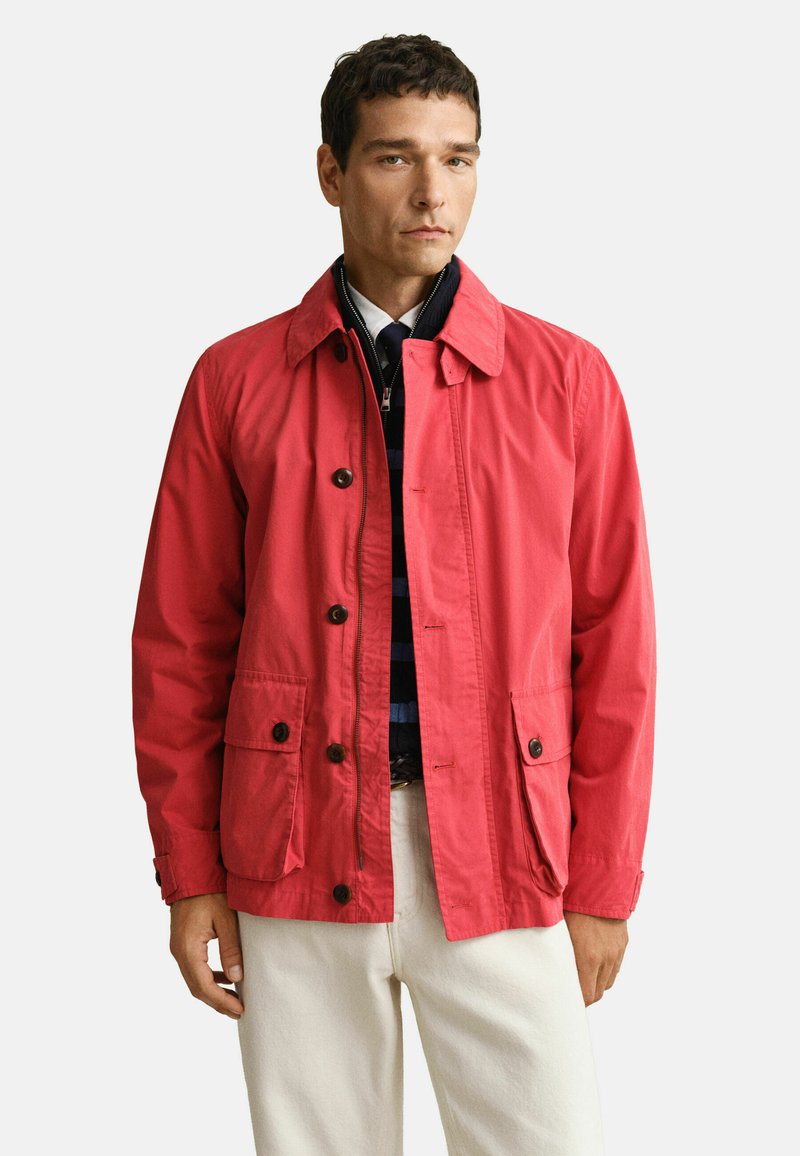 Homme portant une veste corail rouge boutonnée sur un pull zippé bleu marine et un pantalon blanc, debout devant un fond clair uni.