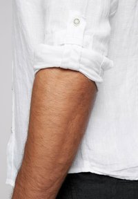 Camisa blanca de lino con mangas remangadas, que presenta un detalle de botón. El vello visible en el brazo contrasta con la tela y textura claras.