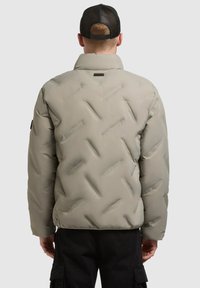 Uomo che indossa una giacca imbottita beige e un cappellino in rete nero, volto di spalle, in piedi contro uno sfondo chiaro e uniforme.