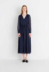 Navy blue maxi dress with long sheer sleeves, V-neckline, and a drawstring waist. Smooth fabric with a flowing silhouette, paired with black shoes.