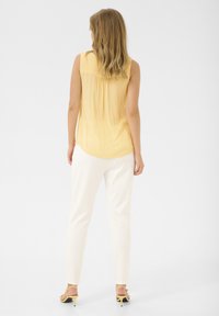 Blusa amarilla sin mangas con una textura suave y pliegues en la parte trasera, combinada con pantalones blancos de corte entallado y sandalias amarillas de tacón.
