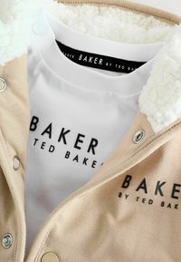 Beige jakk, millel on koorevalge valejõe kaelus, mustade brändingutega valgel siset-särgil, nööbid ja pehme tekstuur.