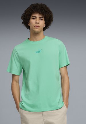 Joven con cabello rizado que lleva una camiseta verde menta de Puma y pantalones beige, de pie con las manos en los bolsillos contra un fondo gris.