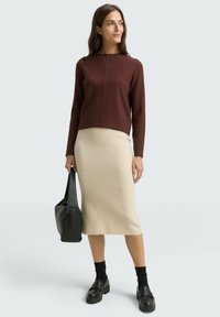 Pull en maille brun à col rond, jupe beige ajustée, chaussettes noires mi-mollet, mocassins noirs épais et sac à main noir.