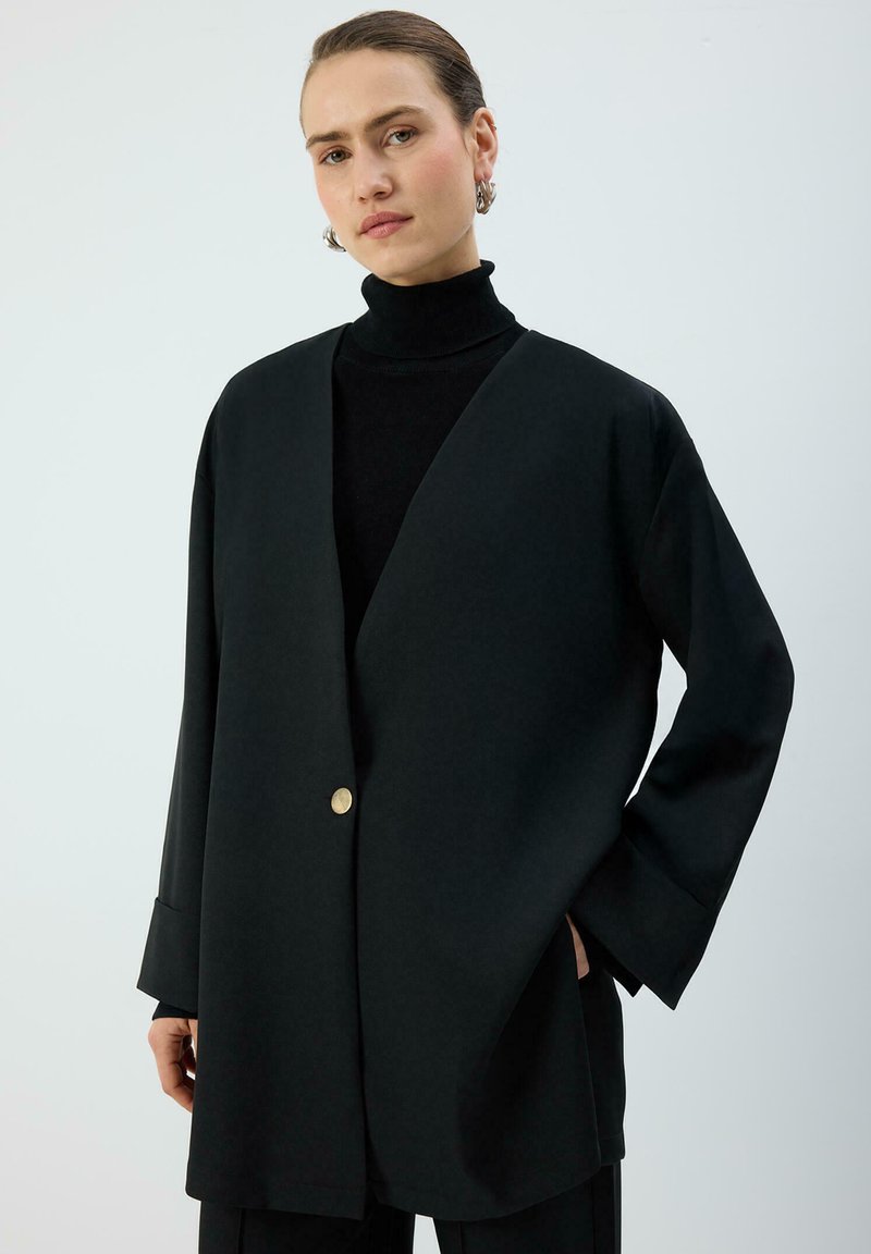 Melns pārmēru lielizmēra žakete ar V veida izgriezumu, viena zelta pogas detaļa, garas piedurknes ar platiem aprocēm, gluda tekstūra, kopā ar augstas apkakles topu.