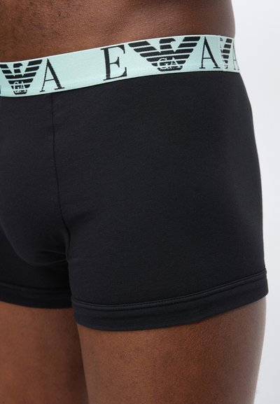 Κοντινό πλάνο προσώπου που φοράει μαύρα ανδρικά εσώρουχα boxer με γαλάζια μέση που έχει επαναλαμβανόμενο μαύρο λογότυπο EA και γράμματα.
