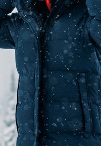 Persona con una chaqueta acolchada azul oscuro con capucha y cremallera, en medio de la nieve que cae, con un fondo nevado desenfocado.