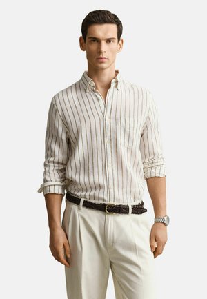 Homme portant une chemise beige à rayures boutonnée avec les manches retroussées, un pantalon beige clair, une ceinture tressée foncée et une montre-bracelet argentée.