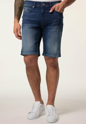 Homme portant un short en jean bleu et des baskets blanches, avec une main dans la poche et un tatouage visible sur l'avant-bras gauche et la cheville droite.