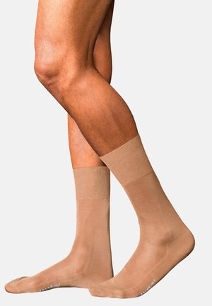 Piernas masculinas con calcetines de compresión beige, cruzadas frente a un fondo claro y liso.