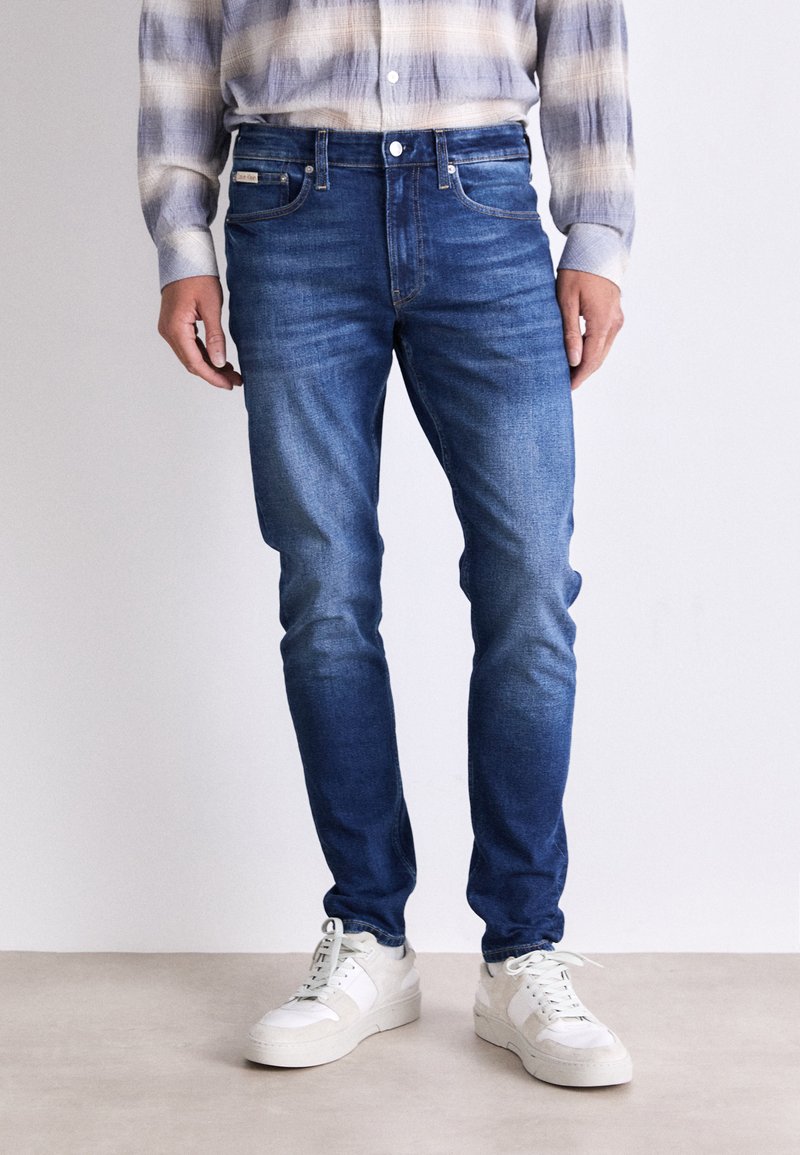 Jeans de denim azul ajustados con un acabado desgastado y un sutil efecto de arrugas. Cuenta con cinco bolsillos y un cierre clásico de botones. Combinados con zapatillas blancas.