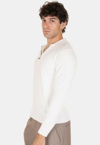 Camicia bianca a maniche lunghe con zip, texture a coste e design aderente, abbinata a pantaloni sartoriali marrone chiaro.