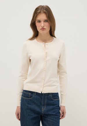 Joven con cabello largo castaño claro que lleva un cárdigan crema abotonado y jeans azules, de pie frente a un fondo claro y liso.