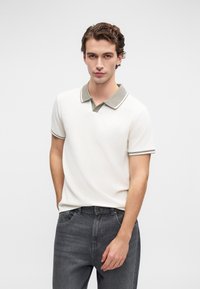 Junger Mann mit welligem braunem Haar, der ein weißes Polohemd mit grauem Kragen und schwarze Jeans trägt, steht vor einem schlichten hellen Hintergrund.