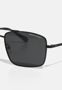 Ochelari de soare rectangulari negri cu lentile întunecate, rame subțiri din metal negru și brand pe lentila din colțul din stânga sus și pe brațul lateral.