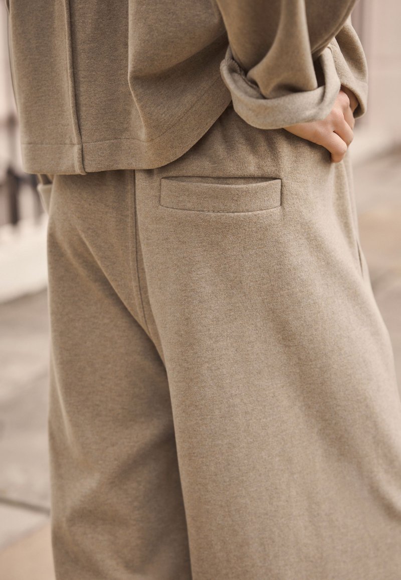 Lähedalt vaadatuna isik, kellel on seljas beežid pehmed riidest püksid ja sobiva värvusega ülaosa, millel on tagatasku ning käsi toetub puusale.