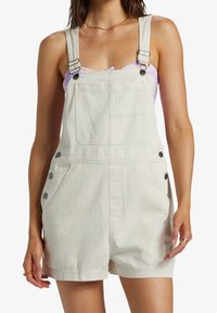Salopette in denim beige con spalline regolabili, due tasche anteriori e dettagli in metallo. Caratterizzata da una vestibilità rilassata di lunghezza ginocchio e tessuto testurizzato.