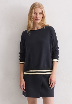 Marineblauer Pullover mit lockerem Schnitt und cremefarbenen Streifen an Bündchen und Saum. Kombiniert mit einem passenden dunkelblauen Rock.