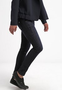 Jeans skinny de mezclilla de lavado oscuro con ligeras marcas de desgaste, combinados con una chaqueta negra. Modelo usando zapatillas negras, de pie en perfil.