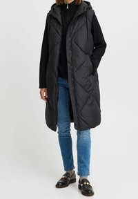 Gestepptes schwarzes Weste mit Kapuze, ausgestattet mit einem Reißverschluss und seitlichen Taschen. Getragen über einem schwarzen Oberteil und blauen Jeans.