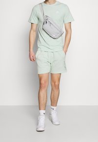 Hellgrünes T-Shirt mit Nike-Logo, passende Shorts, graue Umhängetasche, weiße Sneakers und weiße Socken mit schwarzen Logo-Elementen.