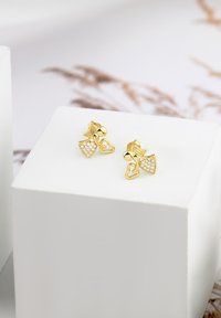 Orecchini dorati con design a cuore e a campana, arricchiti da accenti di cristallo trasparente su una superficie testurizzata, esposti su un piedistallo bianco.