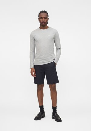 Άνδρας που στέκεται φορώντας ανοιχτό γκρι μακρυμάνικο πουκάμισο, μαύρα σορτς μέχρι το γόνατο, μαύρες κάλτσες και μαύρα loafers σε απλό φόντο.