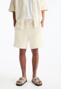 Ensemble coordonné composé d'une chemise et d'un short en texture crémeuse, avec une coupe décontractée et une taille à cordon. Associé à des sandales grises et un T-shirt blanc.