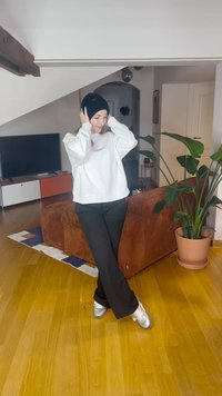 Fehér pulóver, fekete nadrág és fekete sapka. Ezüst sportcipők. Beltéri környezet barna kanapéval és nagy zöld növénnyel.