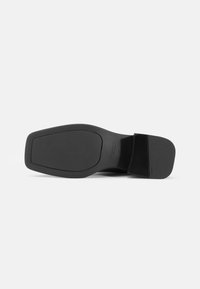 Suola di scarpa nera con superficie liscia e texturizzata. Forma rettangolare, ampia area per le dita e tallone sagomato per la stabilità. Dettaglio del marchio impresso.
