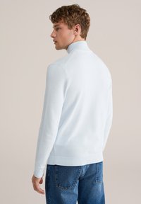 Svetlo modra teksturirana majica z visokim ovratnikom in rebrastimi manšetami, kombinirana z modrimi kavbojkami. Dizajn je oprijet s gladkim zaključkom.