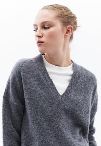 Szary sweter w serek o miękkiej fakturze, noszony na białej koszuli z długim rękawem; ma obniżone ramiona i luźny krój.