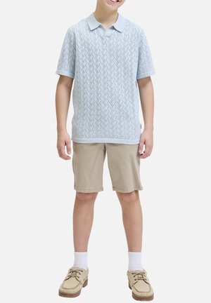Enfant portant un polo tricoté bleu clair à manches courtes, un short beige, des chaussettes blanches et des chaussures à lacets couleur beige, souriant devant un fond uni.