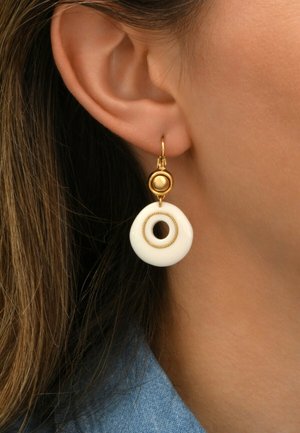 Boucles d'oreilles en métal doré avec un disque circulaire blanc comportant un trou central, rehaussé d'une petite perle dorée ronde au-dessus.