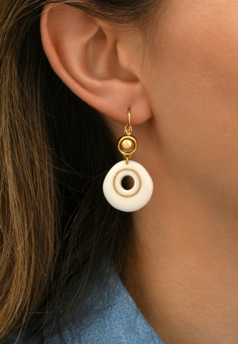 Boucles d'oreilles en métal doré avec un disque circulaire blanc comportant un trou central, rehaussé d'une petite perle dorée ronde au-dessus.