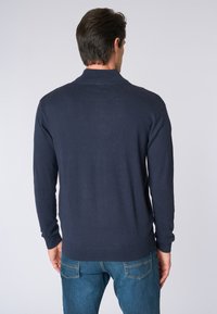Maglione a maniche lunghe di colore blu navy con colletto e polsini a costine, realizzato in un tessuto a maglia liscia, indossato sopra dei jeans blu.