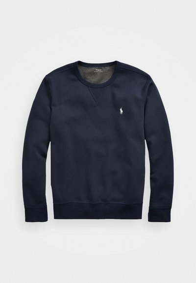 Mornarsko modra trenirka z dolgimi rokavi, krojnim ovratnikom in rebrastimi manšetami. Ima majhen bel logotip na levem prsnem delu in gladko teksturo tkanine.