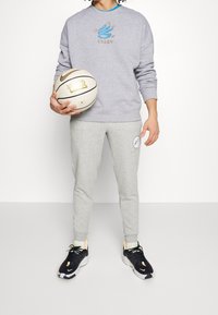 Sweatshirt gris avec le logo "Curry", joggings gris, chaussures de sport noires, et tenant un ballon de basket blanc avec des accents dorés.