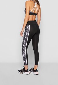 Kobieta stoi w czarnych legginsach sportowych z wzorzystymi bocznymi panelami oraz dopasowanym sportowym stanikiem na ramiączkach, nosząc czarne i różowe buty do biegania.