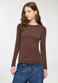 Giovane donna con capelli castani mossi indossa una maglietta marrone a maniche lunghe aderente e pantaloni jeans blu larghi, guardando leggermente verso sinistra.